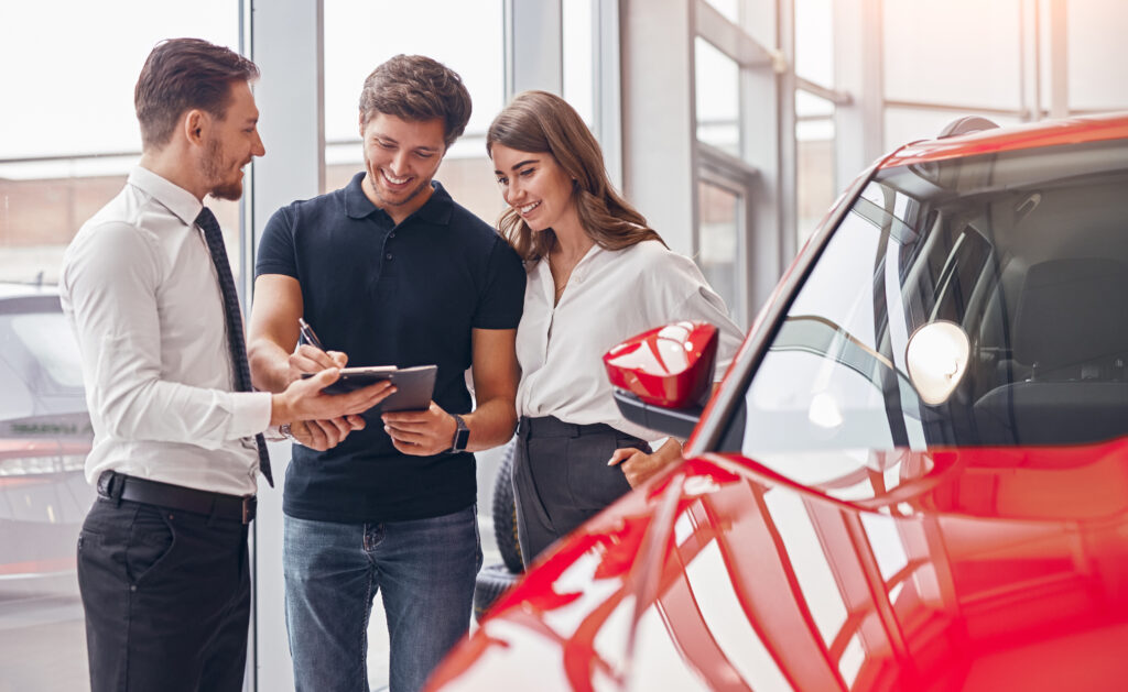 Šťastný mladý muž a žena podepisují smlouvu s prodejcem při koupi nového vozidla v moderním autosalonu.