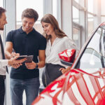 Šťastný mladý muž a žena podepisují smlouvu s prodejcem při koupi nového vozidla v moderním autosalonu.
