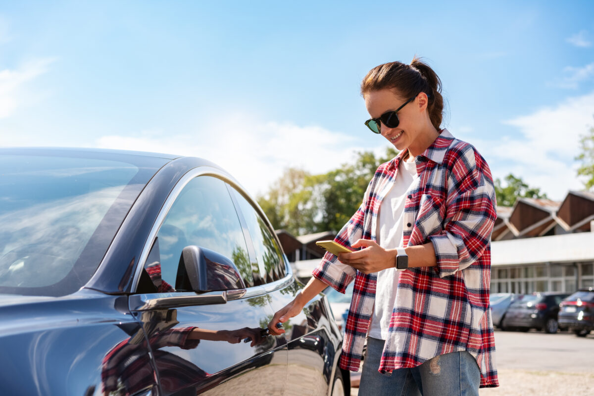 Kavkazská žena v ležérním oblečení odemyká pronajaté auto pomocí svého chytrého telefonu a mobilní aplikace venku v létě.