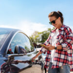 Kavkazská žena v ležérním oblečení odemyká pronajaté auto pomocí svého chytrého telefonu a mobilní aplikace venku v létě.
