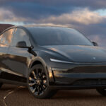 Tesla model Y v tmavě šedé barvě na pískovém podkladu s mraky v pozadí